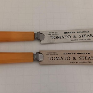 Puede incluir: Dos cuchillos con mangos rectangulares de color naranja y hojas plateadas planas y rectangulares. Las hojas tienen impreso el texto: Henry's Original Tomato & Steak Knife, Surgical Stainless Steel, Henry Fox Solo Distributor, Atlantic City, N.J.
