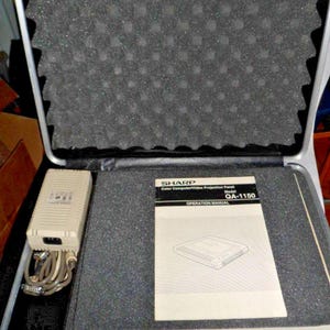 May include: A Sharp QA-1150 Color Computer/Video Projection Panel operation manual, a beige power supply unit with attached cables, and a long beige plastic component, all housed within a black foam-lined carrying case with an egg-crate lid.