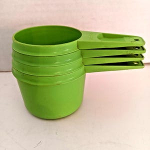 May include: Four green plastic measuring cups stacked together. Each cup has a handle with a hole. The cups are used for measuring ingredients. The cups are in a bright green colour and are nested together.