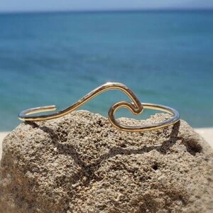 May include: A gold wave-shaped bracelet on a rock with a beach and ocean in the background.