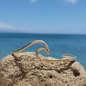 May include: A gold wave-shaped bracelet on a rock with a beach and ocean background.