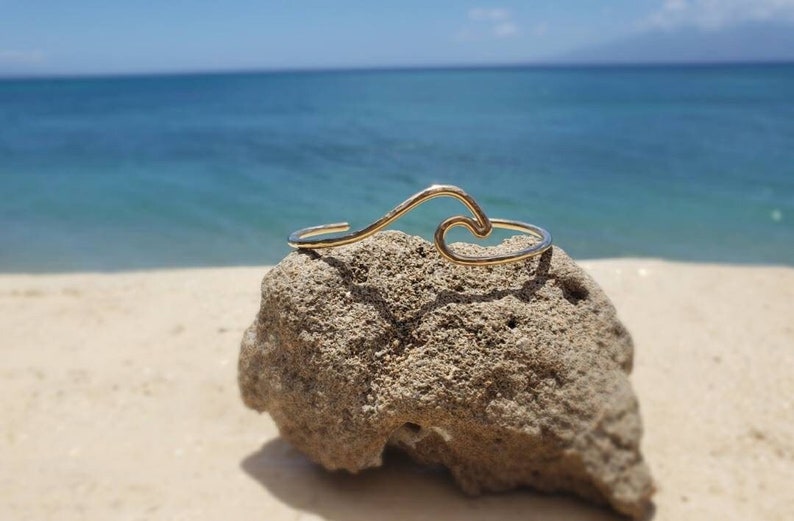 May include: A gold wave-shaped bracelet on a rock with a beach and ocean in the background.