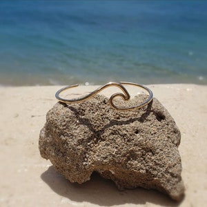 May include: A gold wave-shaped bangle bracelet on a rock with a blurred background of a beach and ocean.