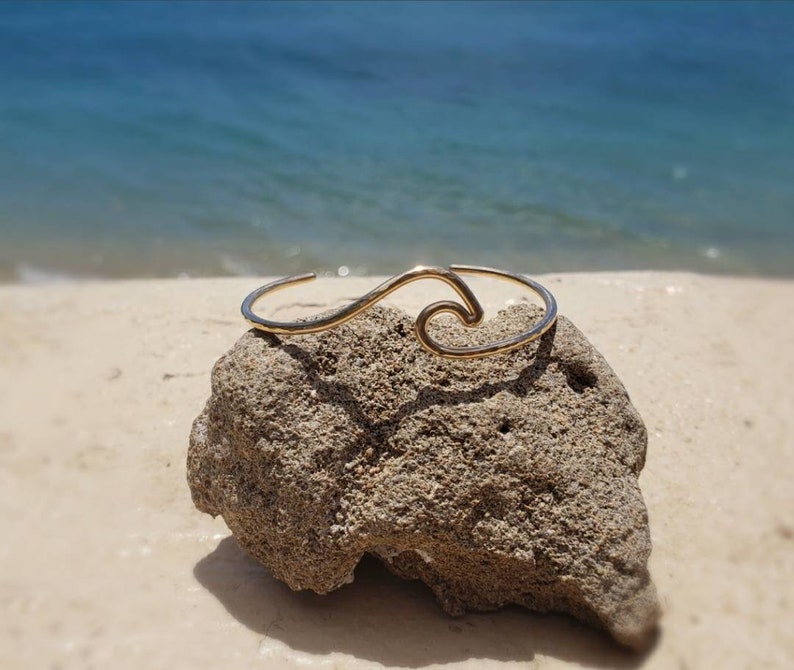 May include: A gold wave-shaped bangle bracelet on a rock with a blurred background of a beach and ocean.