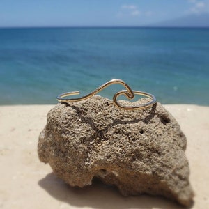 May include: A gold wave-shaped bracelet on a rock with a beach and ocean in the background.