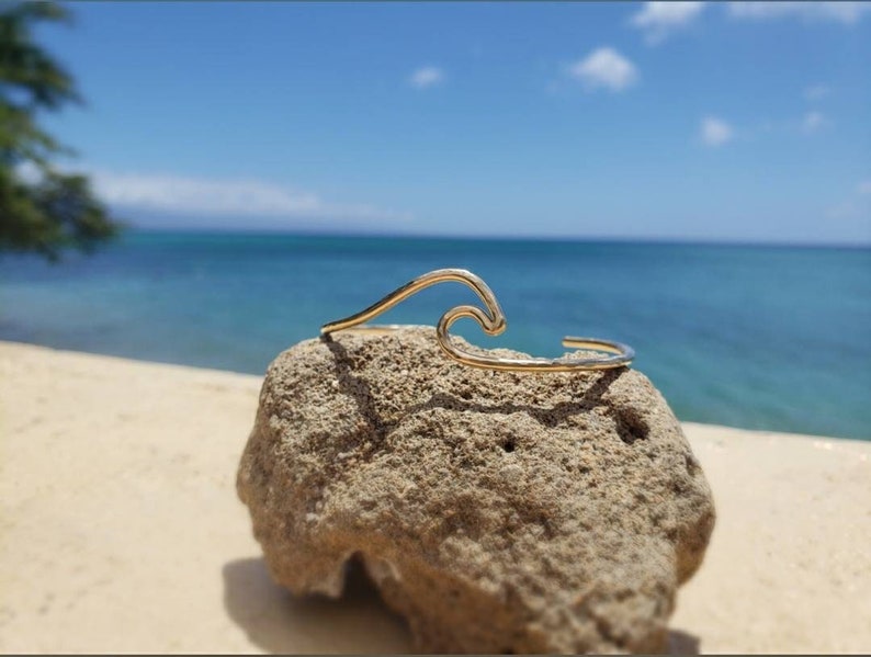 May include: A gold wave-shaped bracelet on a rock with a beach and ocean background.