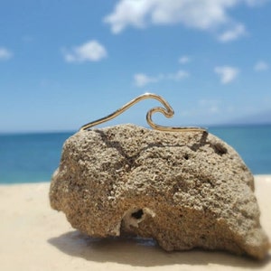 May include: A gold wave-shaped wire bracelet on a rock with a beach and ocean background.