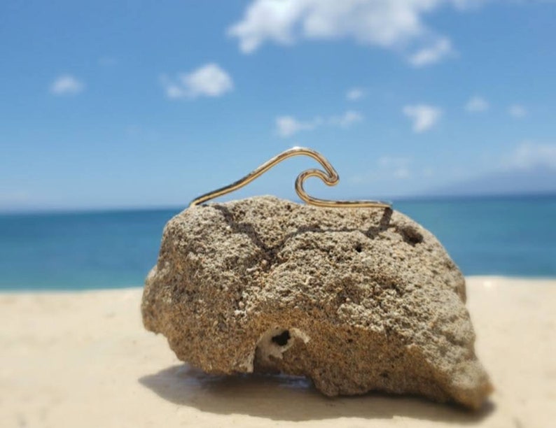 May include: A gold wave-shaped wire bracelet on a rock with a beach and ocean background.