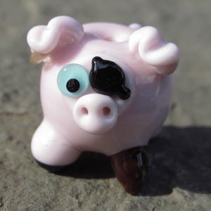 May include: A small, pink glass pig figurine with a light blue eye and a black eye patch. The pig has curled ears, a small snout, and brown feet. This decorative item is a charming collectible.