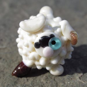 May include: A whimsical, handcrafted glass sheep figurine. The sheep features a white, textured body, a black and white face, and a light blue eye. The sheep's hoof is brown, and its ear has a gold glitter accent.