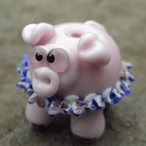 May include: A pink glass pig ornament with big eyes and a blue and white frilled collar. The pig has a round body, small legs, and curled ears. A decorative and whimsical item.