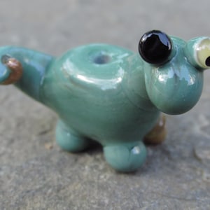 May include: A whimsical, teal-colored glass figurine shaped like a small, stylized creature. It features large, expressive eyes, a rounded body, and small legs. The figurine has a hole on top and a copper-colored detail.