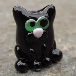 May include: A glossy black cat figurine with large green eyes and a pink nose. The cat is sitting, with its paws and ears visible. The figurine has a smooth, reflective surface and a whimsical design.