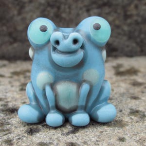 May include: A whimsical, light blue frog figurine with large, round eyes and a smiling expression. The frog has light blue spots and is made of a smooth, possibly glass-like material. The frog is sitting on a stone surface.