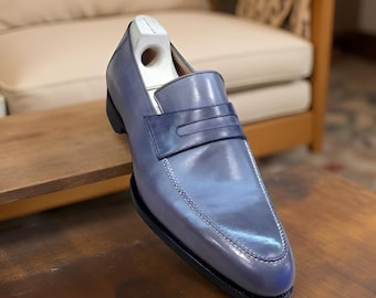 May include: A blue leather loafer with a dark blue accent on the vamp. The shoe has a black sole and a wooden shoe tree inside. The leather has a polished finish and visible stitching.