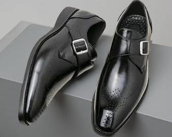 May include: A pair of black leather monk strap shoes with a glossy finish. Each shoe features a silver-colored buckle and decorative perforations on the toe. The shoes are displayed on a gray surface.