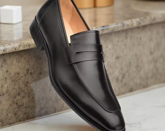 Könnte beinhalten: Schwarzer Leder-Loafer mit eckiger Spitze und Riemen über dem Spann. Der Schuh hat einen niedrigen Absatz und eine glatte, polierte Oberfläche. Die Innenseite ist hellbraun.