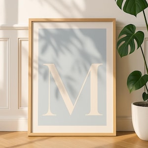 May include: A framed print displaying the letter 'M' in a beige tone against a pale blue backdrop. The artwork is presented within a wooden frame, offering a minimalist design. A potted plant is visible in the background.