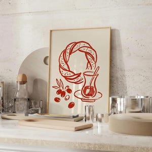 May include: A framed print featuring a red illustration of a bagel and a glass of tea, with olives. The image is set on a white countertop with wooden cutting boards, glass bottles, and metal cups. The overall theme is food and drink.