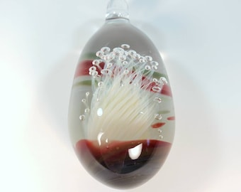 May include: A glass pendant with a white flower-like design inside. The glass is clear and has red, green, and white stripes. There are small air bubbles throughout the glass.