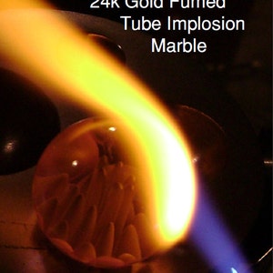 Puede incluir: Un mármol de vidrio con un interior dorado está siendo calentado con un soplete. La llama es de color naranja y amarillo brillante, y el mármol está brillando de rojo. El texto "24k Gold Fumed Tube Implosion Marble" es visible sobre el mármol.