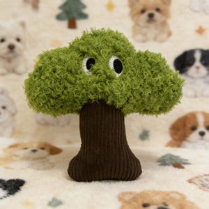 May include: A plush toy shaped like a tree with a green, textured canopy and a brown trunk. The tree has embroidered eyes. The background features a patterned fabric with images of puppies.