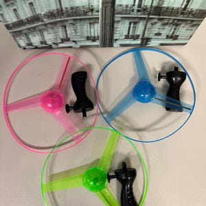 May include: Three colorful flying disc toys. One is pink, one is blue, and one is green. Each disc has three blades and a black handle. The toys are designed for outdoor play and are suitable for all ages.