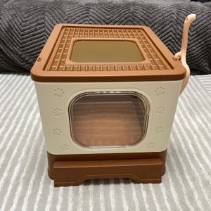 May include: A two-tone cat litter box in cream and brown, featuring a rectangular design with a screened top and a clear front door. Paw prints decorate the cream-colored sides. A matching scoop is attached to the side.