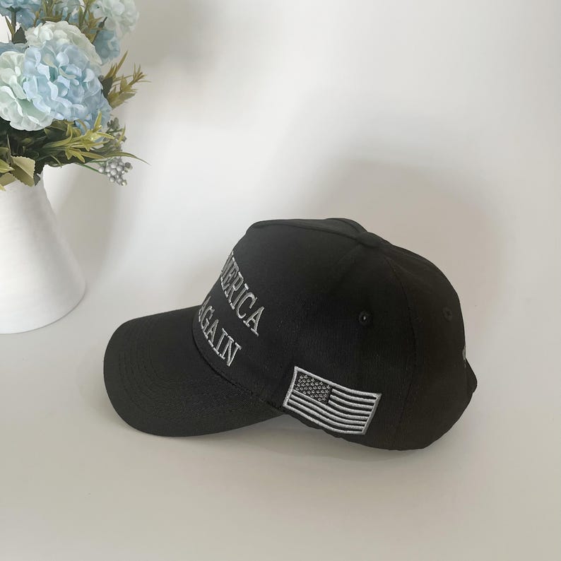 May include: Black baseball cap with the words "AMERICA AGAIN" embroidered in white. An American flag patch is on the side. The cap is made of a durable fabric and has a curved brim.