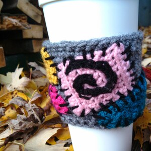 Puede incluir: Una taza de caf&eacute; blanca con un acogedor de punto de crochet gris, rosa, negro, amarillo y azul. El acogedor tiene un dise&ntilde;o de patchwork con un patr&oacute;n en espiral.