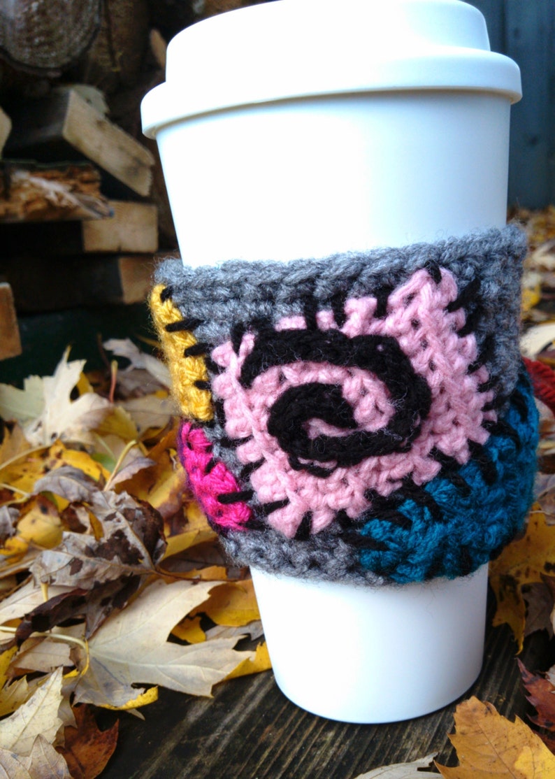 Puede incluir: Una taza de caf&eacute; blanca con un acogedor de punto de crochet gris, rosa, negro, amarillo y azul. El acogedor tiene un dise&ntilde;o de patchwork con un patr&oacute;n en espiral.