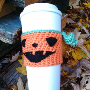 Puede incluir: Una taza de caf&eacute; desechable blanca con una funda de punto naranja con una cara de calabaza negra. La funda tiene un ribete turquesa y una borla turquesa. La taza est&aacute; rodeada de hojas de oto&ntilde;o.