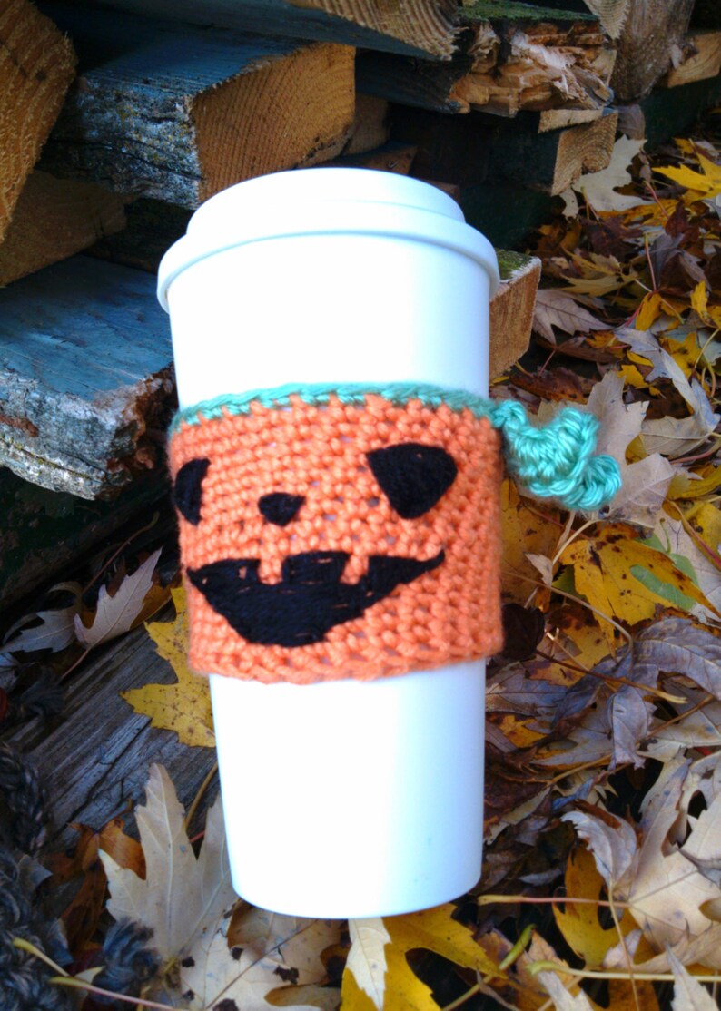 Puede incluir: Una taza de caf&eacute; desechable blanca con una funda de punto naranja con una cara de calabaza negra. La funda tiene un ribete turquesa y una borla turquesa. La taza est&aacute; rodeada de hojas de oto&ntilde;o.