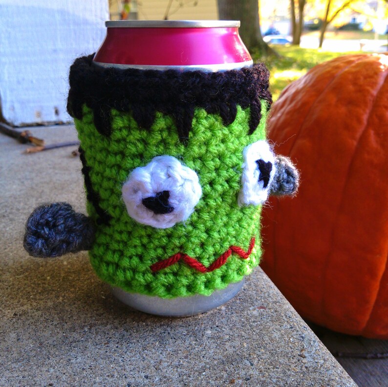 May include: A green crocheted can cozy in the shape of Frankenstein's monster. The cozy has white and black accents for the eyes and hair, a red stitched mouth, and a gray arm. A pink can is visible inside the cozy. A large orange pumpkin is in the background.