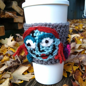 Puede incluir: Un calentador de taza de caf&eacute; de ganchillo con un personaje azul y rojo con ojos grandes. El personaje es una versi&oacute;n estilizada de Sally de la pel&iacute;cula El extra&ntilde;o mundo de Jack.