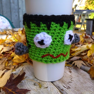 May include: A white disposable coffee cup with a green crocheted Frankenstein monster cozy. The cozy has black hair, white eyes with black pupils, gray bolts, and a red stitched mouth. The cup is on a weathered wooden surface.