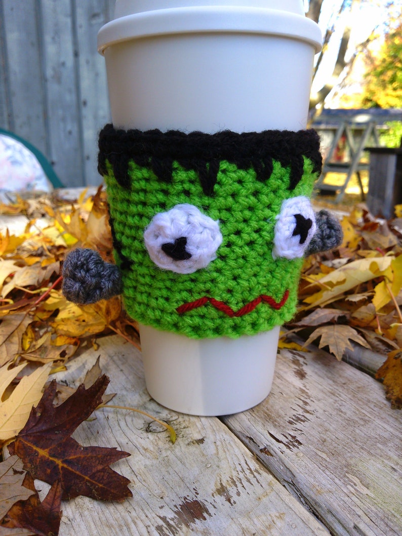 May include: A white disposable coffee cup with a green crocheted Frankenstein monster cozy. The cozy has black hair, white eyes with black pupils, gray bolts, and a red stitched mouth. The cup is on a weathered wooden surface.
