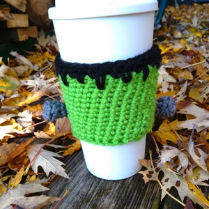 May include: A white disposable coffee cup with a green and black crocheted monster-themed cozy. The cozy has a green body, black hair, and gray pom-pom bolts. The cup is sitting on a wooden surface with fall leaves.