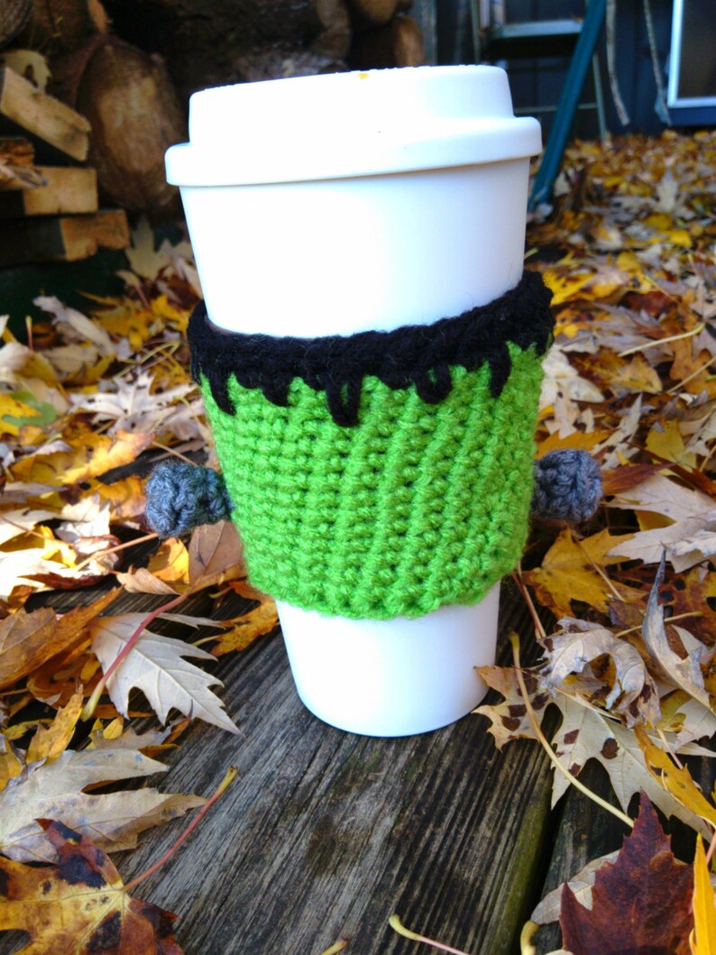 May include: A white disposable coffee cup with a green and black crocheted monster-themed cozy. The cozy has a green body, black hair, and gray pom-pom bolts. The cup is sitting on a wooden surface with fall leaves.