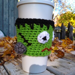 May include: A white travel mug with a green crocheted Frankenstein-themed cozy. The cozy features black hair, a stitched mouth, and a gray stitched bump. The cozy is designed to fit around the mug, providing insulation and a festive Halloween look.