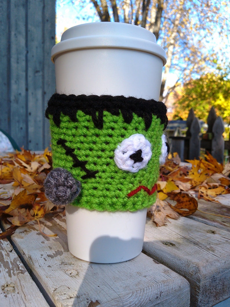 May include: A white travel mug with a green crocheted Frankenstein-themed cozy. The cozy features black hair, a stitched mouth, and a gray stitched bump. The cozy is designed to fit around the mug, providing insulation and a festive Halloween look.
