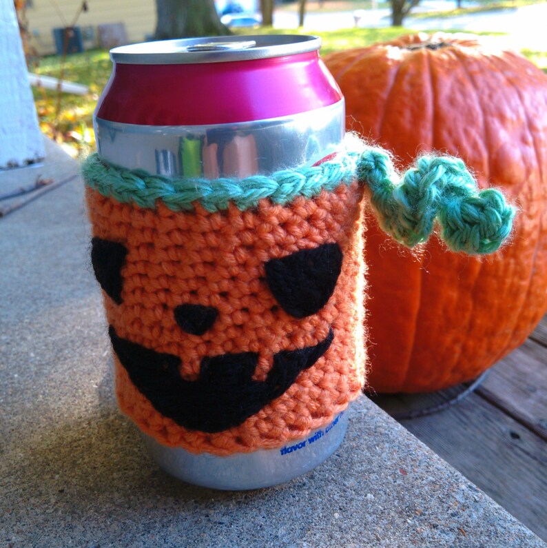 Puede incluir: Un portabotellas de ganchillo naranja hecho a mano con una cara de calabaza, ojos y boca negros y un ribete verde. Se adjunta un asa verde en forma de vid. Una gran calabaza naranja est&aacute; en el fondo.