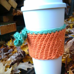 Puede incluir: Una taza de caf&eacute; desechable blanca con una funda naranja tejida y una cinta verde. La funda est&aacute; envuelta alrededor de la taza y est&aacute; hecha de un material texturizado tejido a mano. El fondo incluye hojas de oto&ntilde;o y madera.