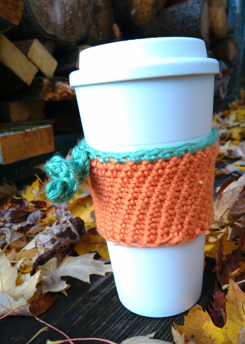 Puede incluir: Una taza de caf&eacute; desechable blanca con una funda naranja tejida y una cinta verde. La funda est&aacute; envuelta alrededor de la taza y est&aacute; hecha de un material texturizado tejido a mano. El fondo incluye hojas de oto&ntilde;o y madera.