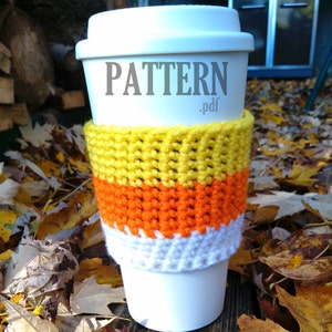 Op de afbeelding: Een witte koffiebeker met een gehaakte candy corn-kleurige cozy. Op de beker staat de tekst "PATTERN .pdf" gedrukt.