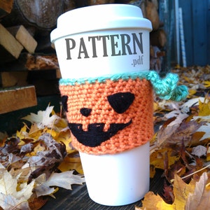 May include: A white coffee cup with a knitted orange pumpkin cozy. The cozy has a black jack-o'-lantern face and a green trim. The word "PATTERN.pdf" is printed on the cup. The cup is surrounded by fall leaves.