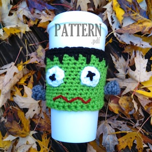 May include: A white disposable coffee cup with a green crocheted Frankenstein monster cozy. The cozy has black hair, white eyes, a red mouth, and gray arms. The word "PATTERN.pdf" is printed on the cup.