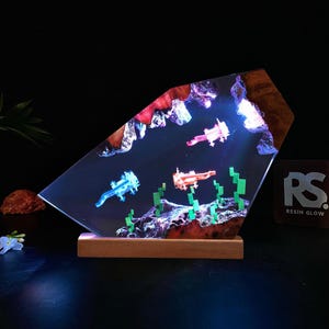 May include: A unique resin art piece featuring three glowing axolotls in a simulated underwater scene. The artwork is set within a clear, angled resin block, with a wooden base and a dark background. The axolotls are in shades of blue, orange, and pink.