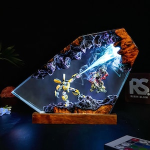May include: A unique resin art piece featuring two robot figures engaged in battle. The artwork is encased in a clear, angled resin block with a wooden base. The resin has a wood grain pattern and a blue light effect.