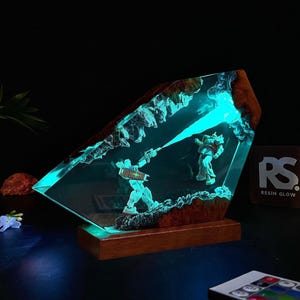 May include: A resin art piece featuring two robot figures in a battle scene, illuminated by a vibrant blue light. The artwork is encased in a clear resin block with a wooden base, creating a unique and eye-catching display.